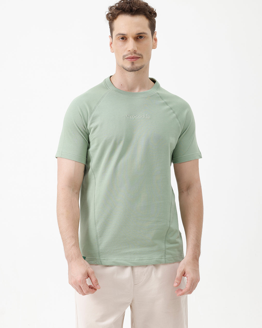 A man wearing a light sage green athletic t-shirt with a small tonal logo on the chest, paired with beige pants, standing against a plain white studio background.