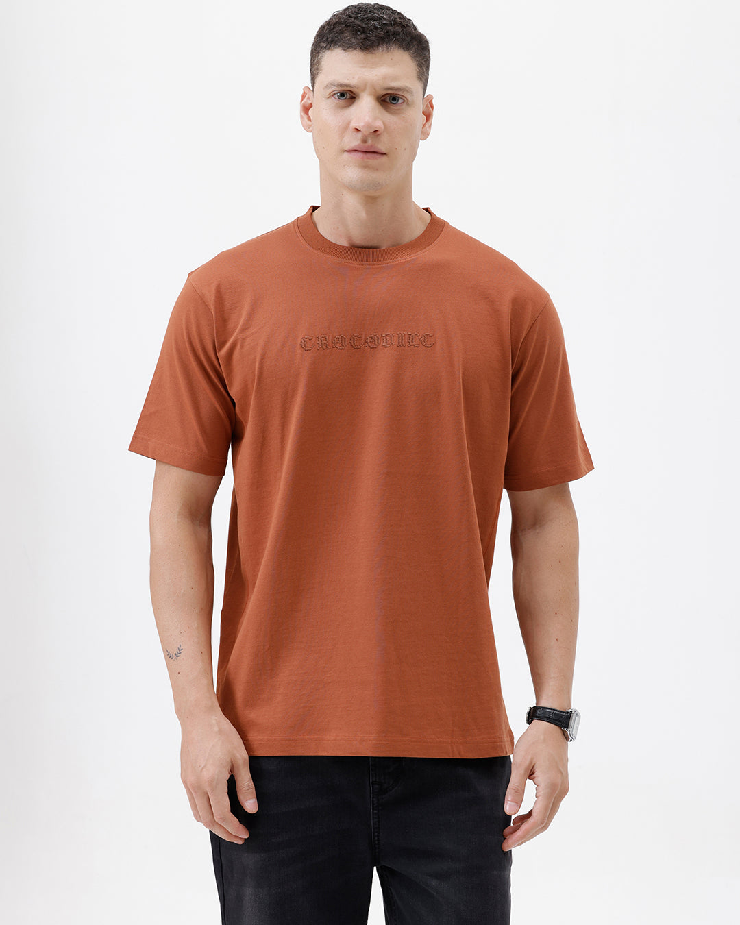 A man wearing a rust-orange crewneck t-shirt with a subtle tonal 'ELECTRIC' logo across the chest, paired with black pants, photographed against a clean white studio background.