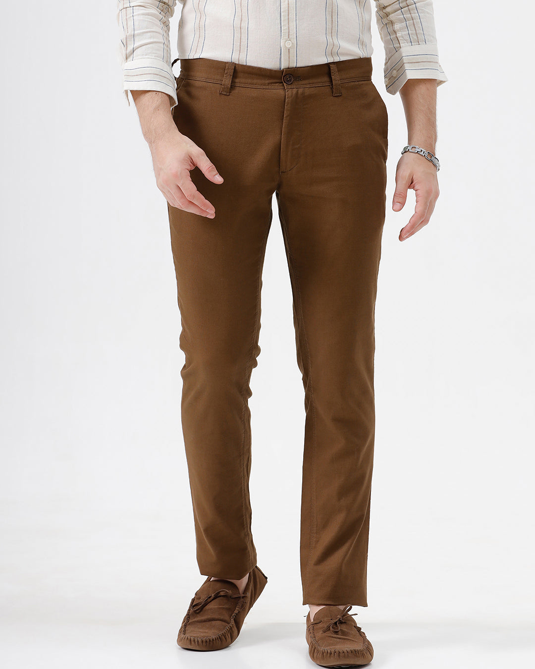 A man wearing brown slim-fit trousers, a cream and brown striped long-sleeve shirt, and brown suede loafers, standing against a plain white studio background.