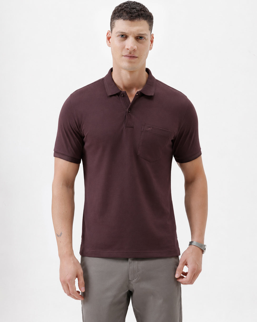 A man wearing a short-sleeve polo shirt in dark burgundy with a chest pocket, paired with gray trousers, standing against a plain light gray background.
