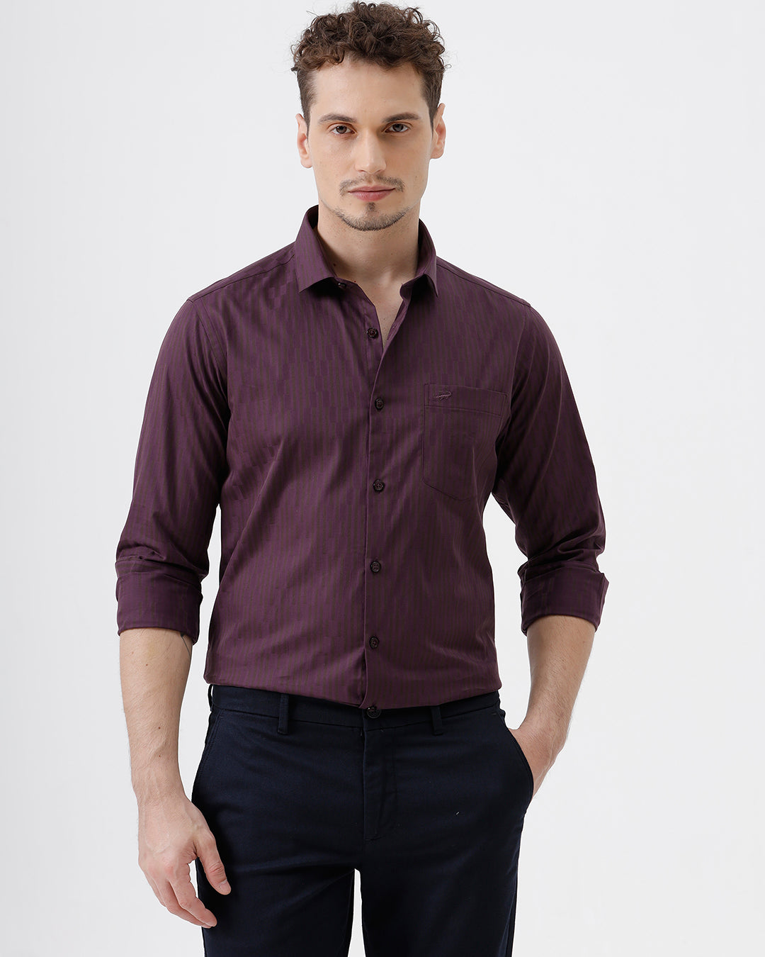 A man wearing a dark purple long-sleeve button-down shirt with subtle vertical stripes and dark navy trousers, standing against a light gray background.