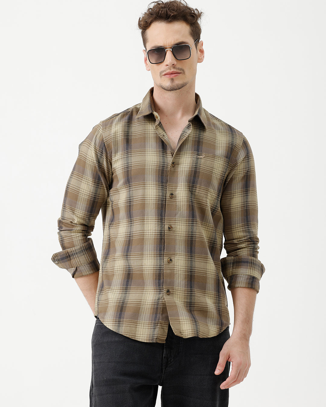 A man wearing a brown and beige plaid long-sleeve button-down shirt with dark sunglasses and black jeans, standing against a plain white studio background.