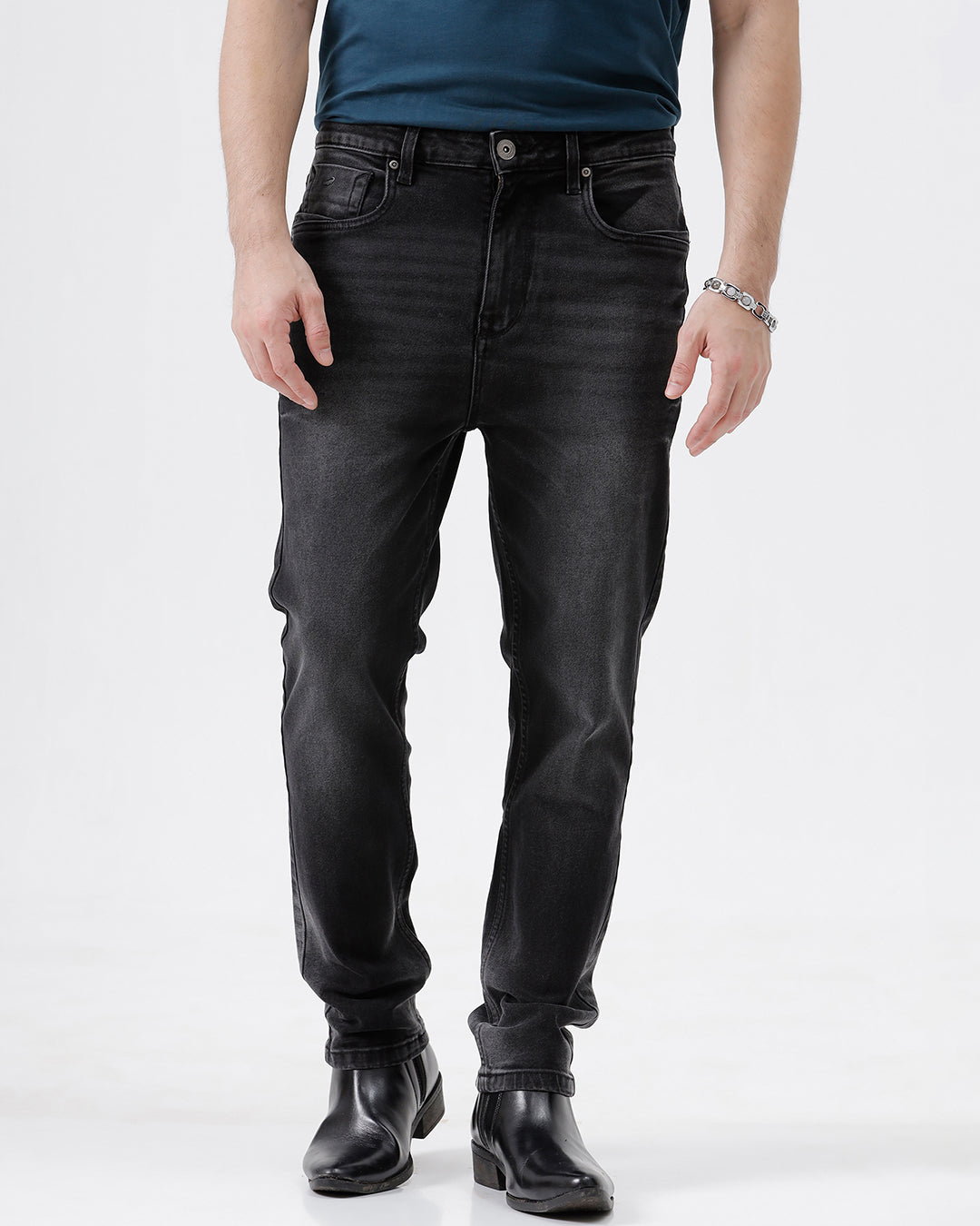 A person wearing slim-fit black denim jeans with a faded wash, paired with a dark blue t-shirt and black leather boots, shown from the waist down on a light gray studio background.
