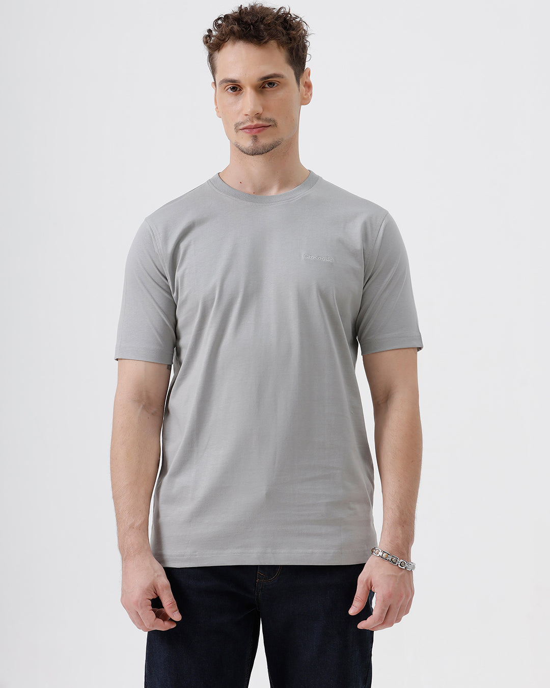 A man wearing a light gray short-sleeve crewneck t-shirt with a subtle tonal chest logo, paired with dark jeans, standing against a light gray studio background.
