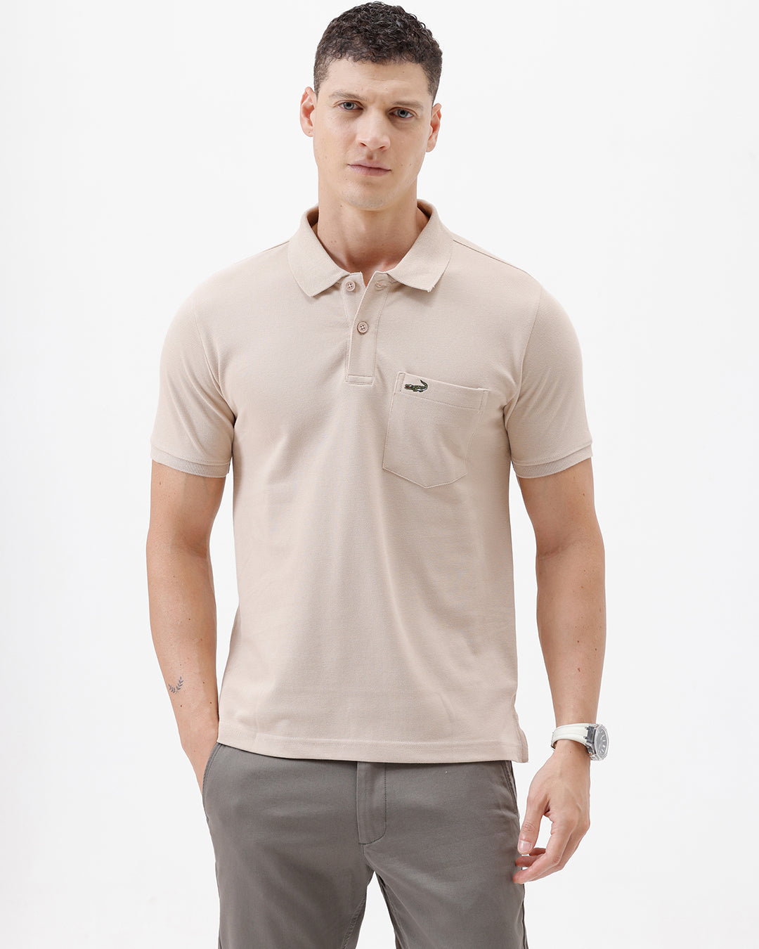 A man wearing a light beige short-sleeve polo shirt with a small crocodile logo on the chest pocket, paired with gray trousers, photographed against a plain white studio background.