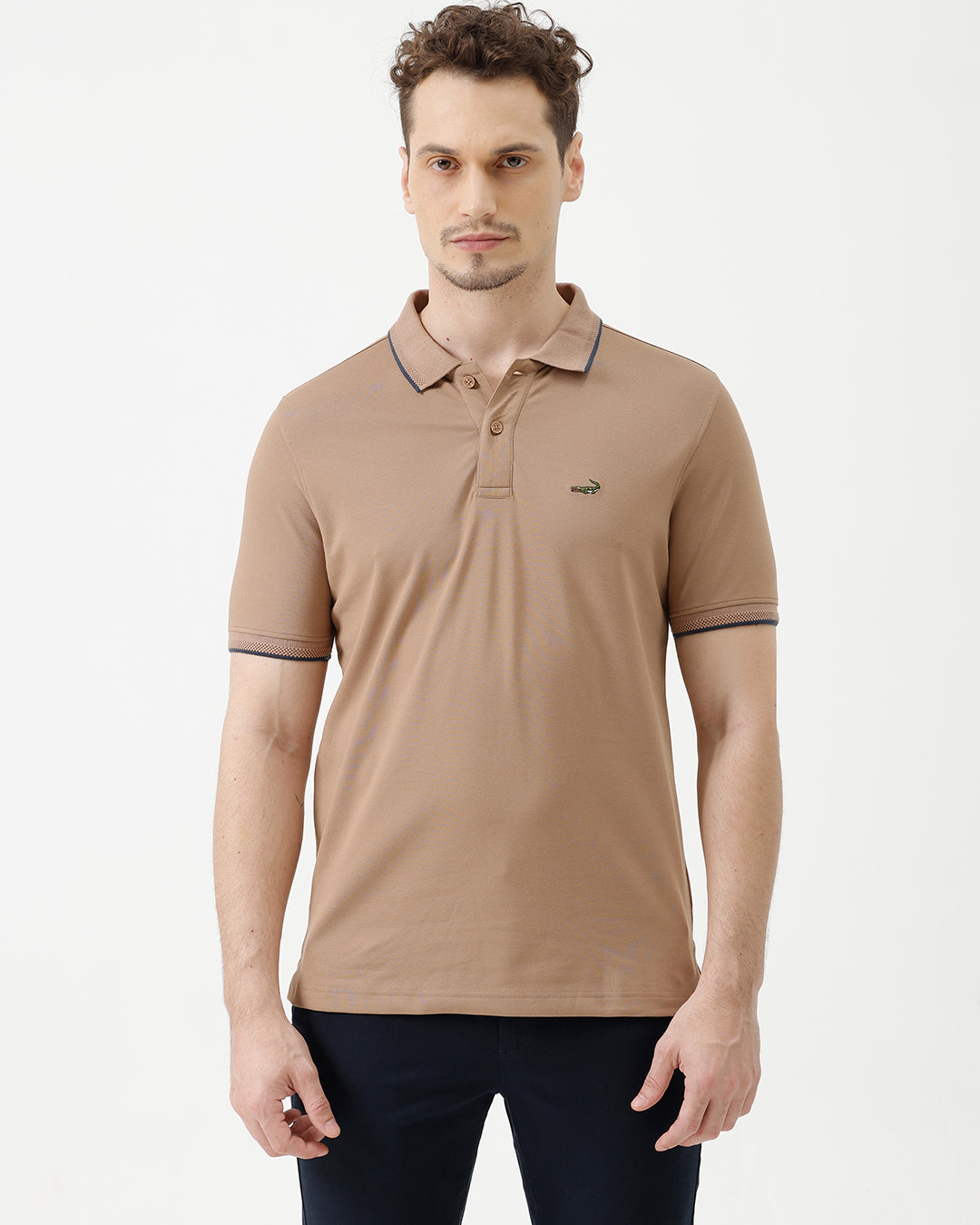 A man wearing a tan short-sleeve polo shirt with dark blue piping on the collar and sleeve cuffs, paired with dark navy pants, against a plain light gray background.