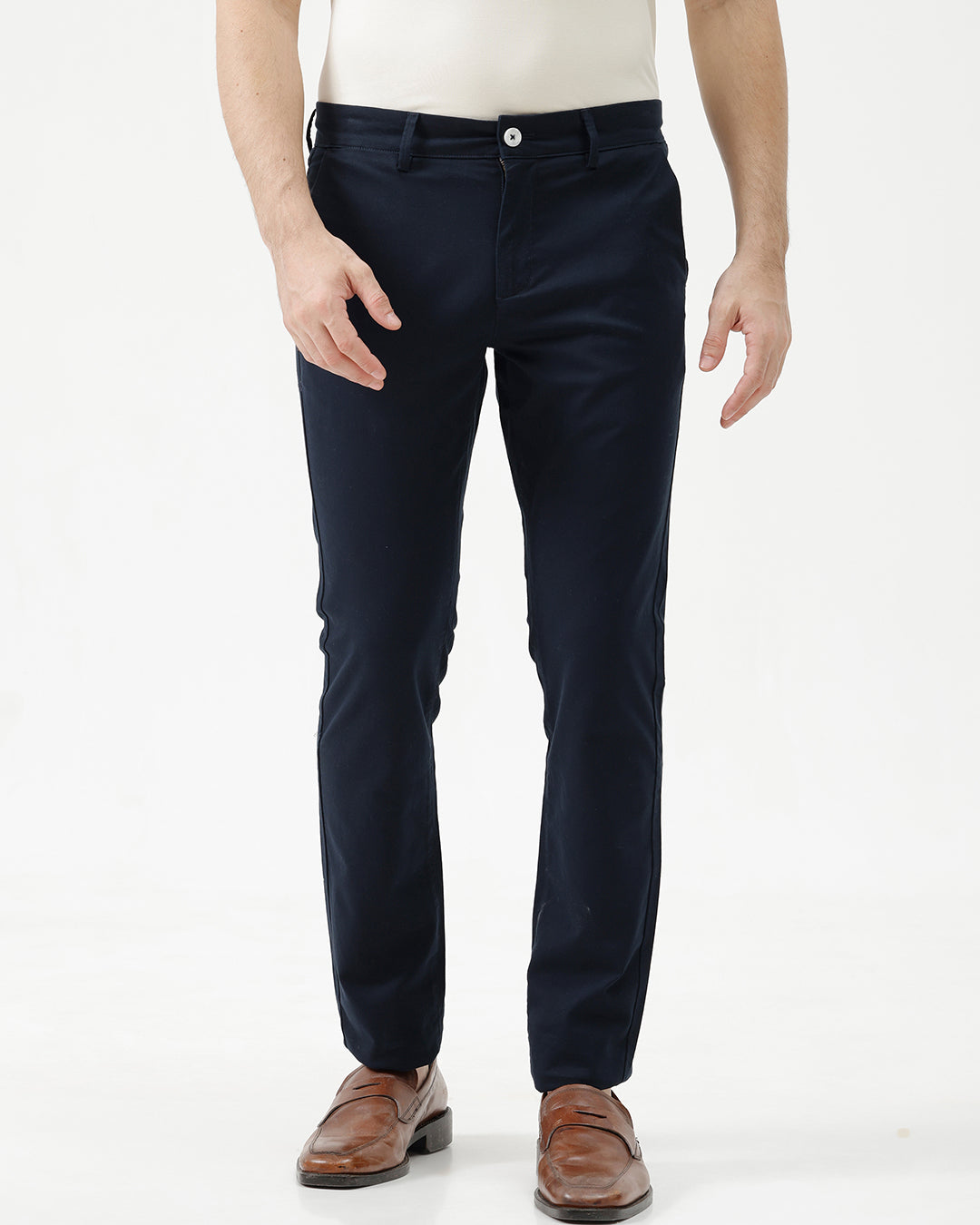 A man wearing dark navy blue slim-fit trousers, a cream-colored shirt, and brown leather loafers, standing against a plain white studio background.