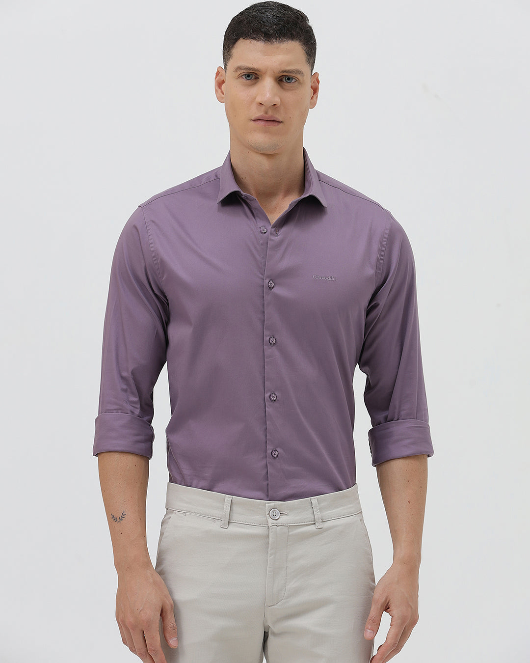 A man wearing a purple long-sleeve button-down shirt with rolled-up sleeves and light beige trousers, standing against a plain light gray background.