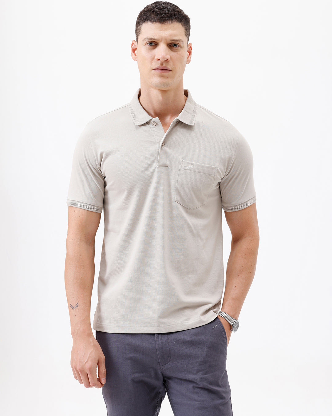 A man wearing a light beige short-sleeve polo shirt with a chest pocket, paired with dark gray trousers, standing against a plain white studio background.