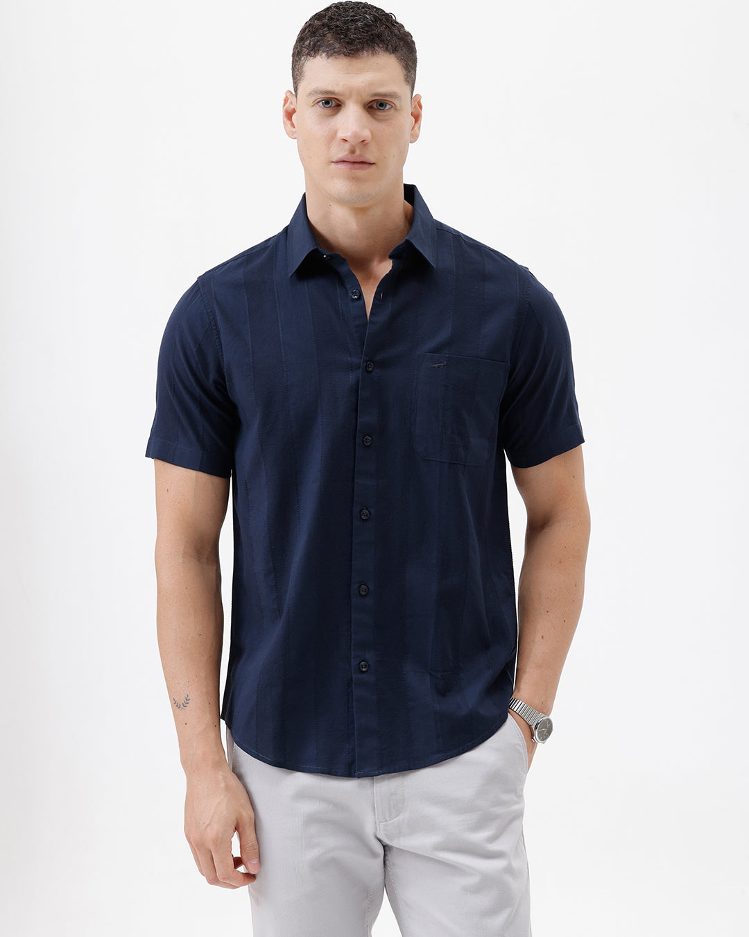 A man wearing a dark navy blue short-sleeve button-down shirt and light gray trousers, standing against a plain white studio background.