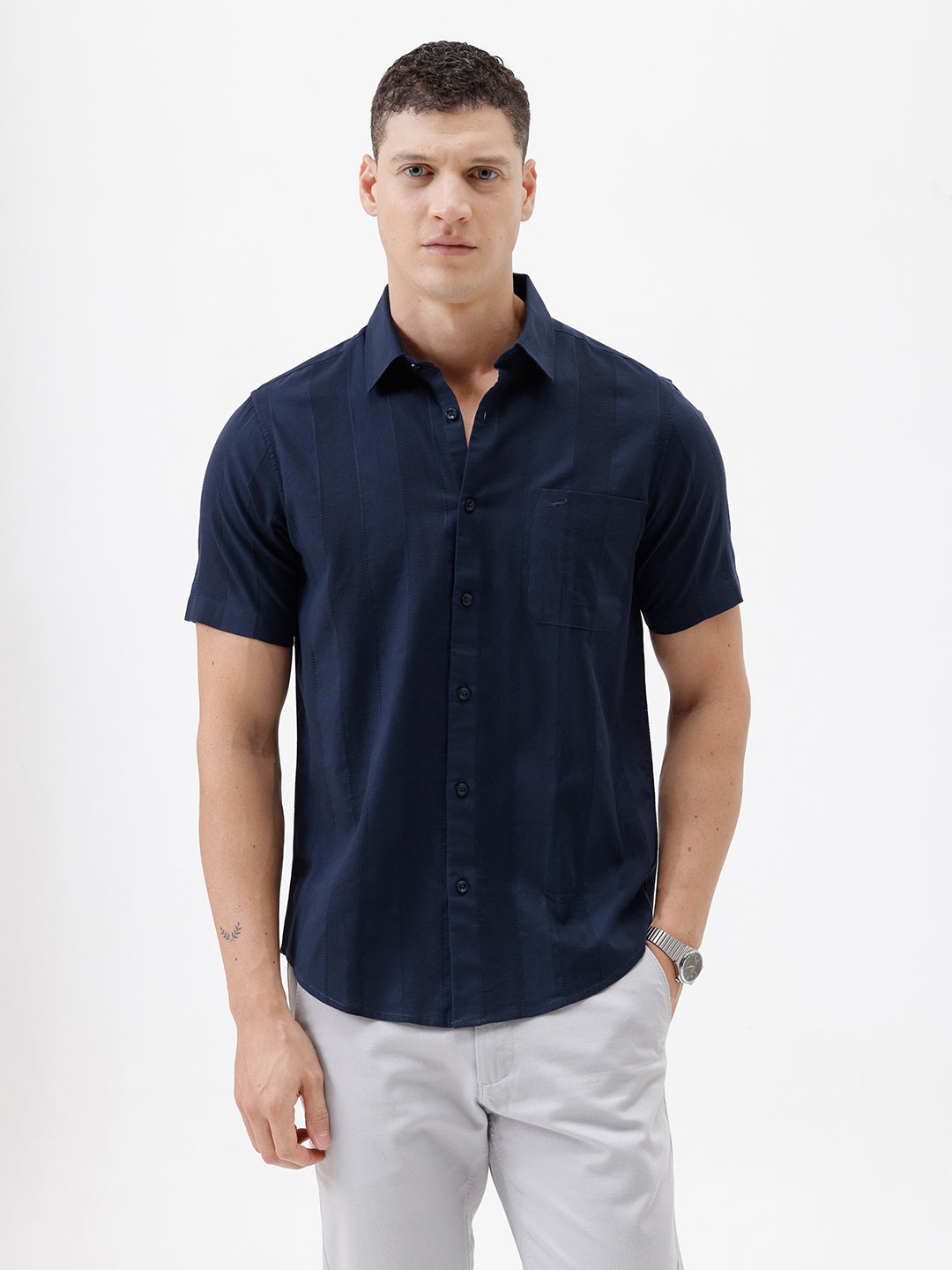 A man wearing a dark navy blue short-sleeve button-down shirt and light gray trousers, standing against a plain white studio background.