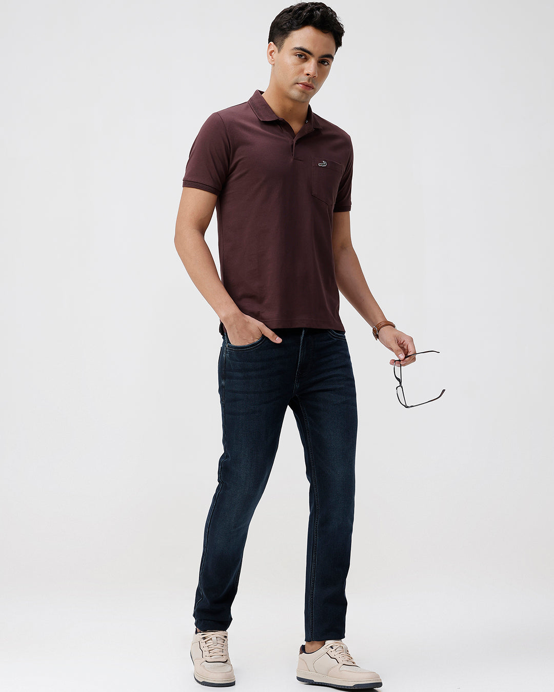 Man wearing a brown polo shirt and dark jeans holding eyeglasses on a white background