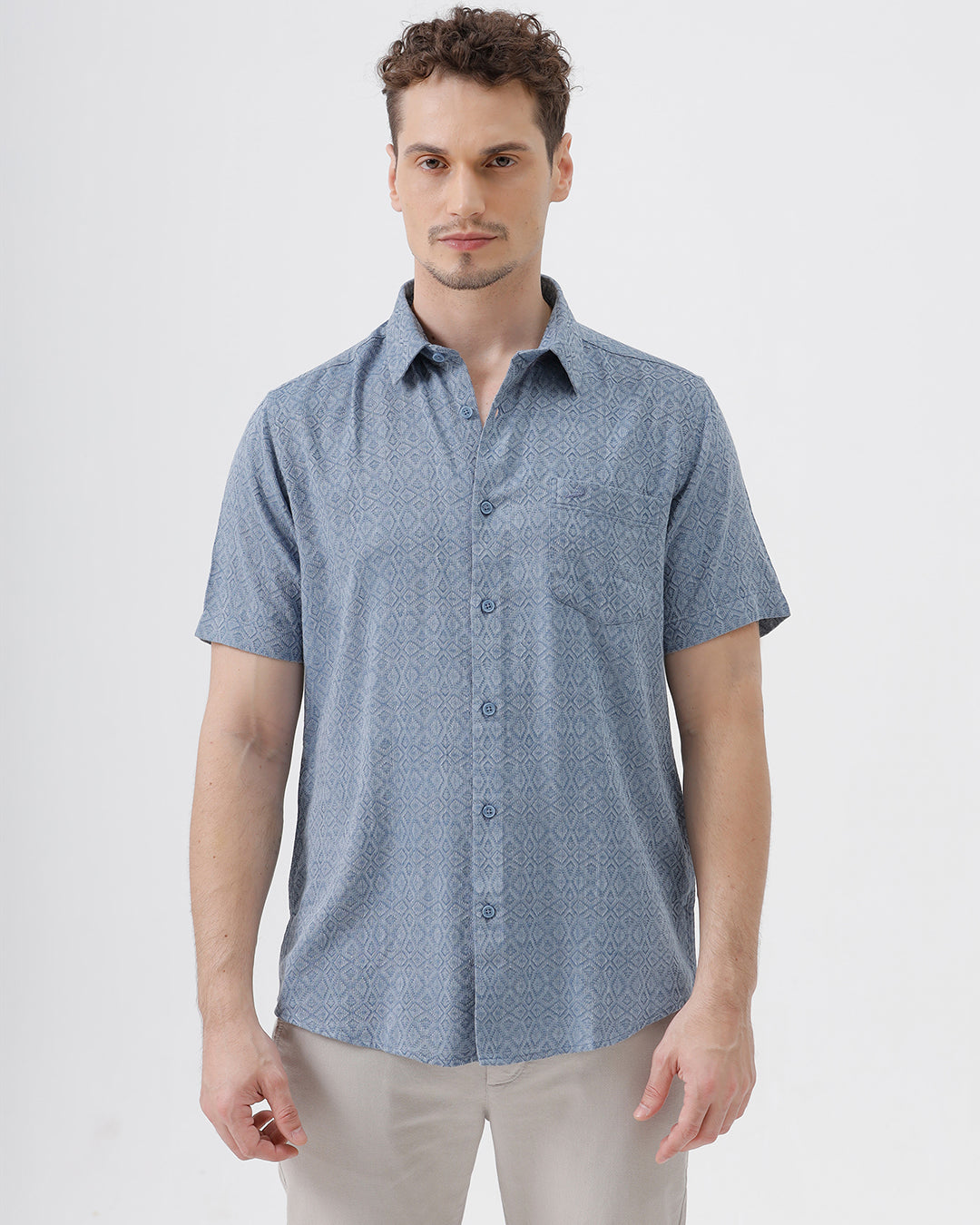 A man wearing a short-sleeve button-up shirt with a fine blue and white geometric pattern, paired with light beige trousers, standing against a plain light gray background.