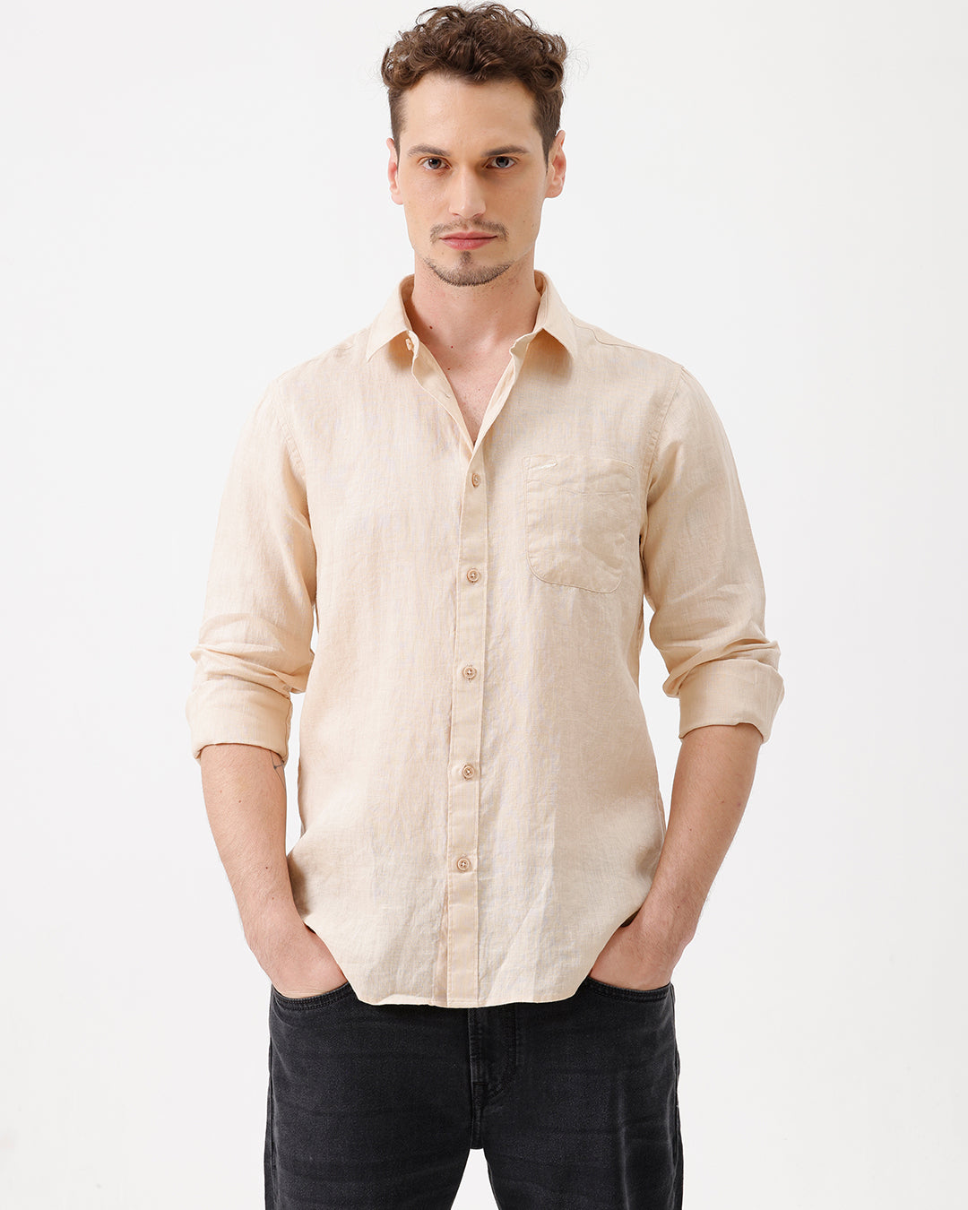 A man wearing a light beige linen button-up shirt with rolled-up sleeves and dark jeans, standing against a plain white studio background.
