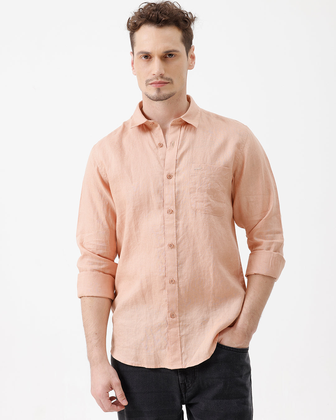 A man wearing a light peach-colored linen button-down shirt with dark jeans, standing against a plain white studio background.