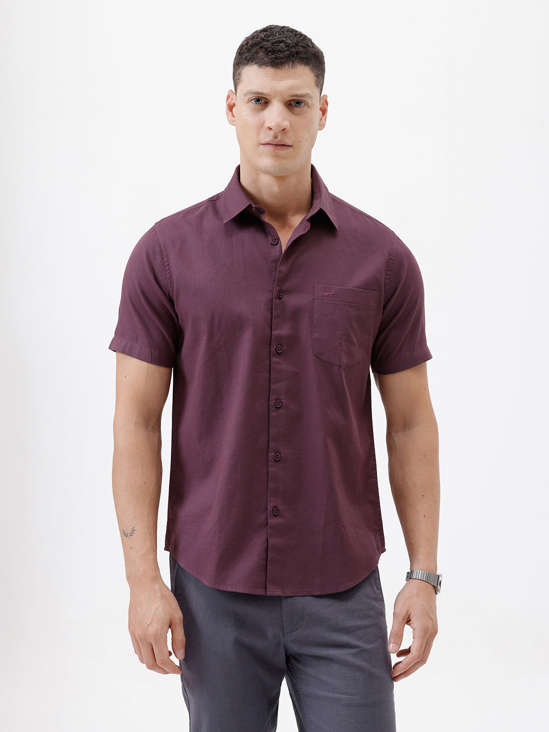 A man modeling a short-sleeve button-up shirt in a deep plum purple color, paired with gray trousers, against a plain white studio background.