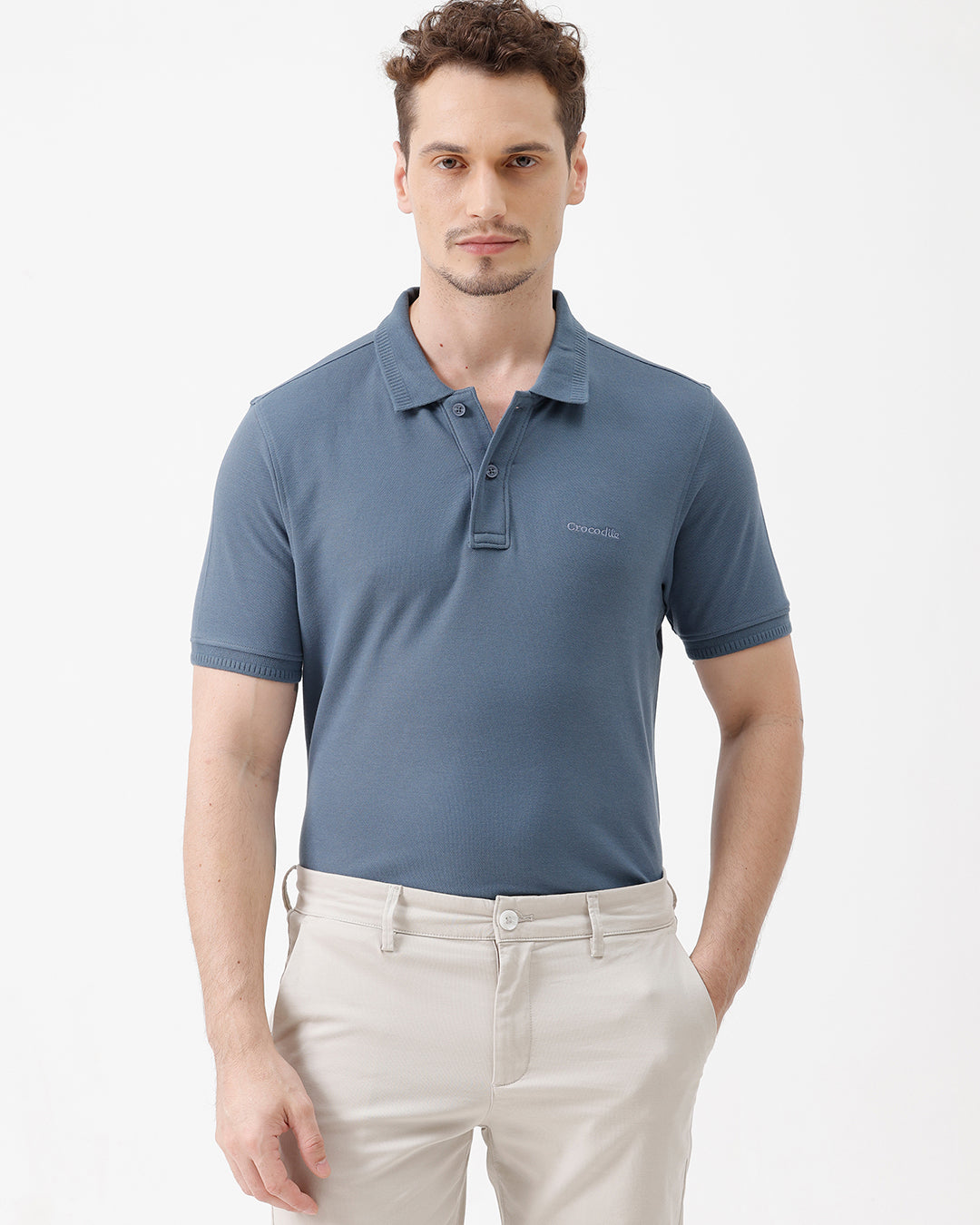 A man wearing a slate blue short-sleeve polo shirt and beige trousers, standing against a plain white studio background.