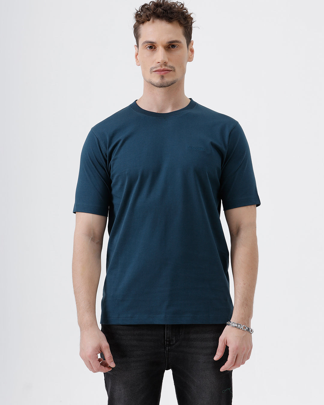 A man wearing a dark navy blue crew-neck t-shirt and black jeans, standing against a light gray studio background.