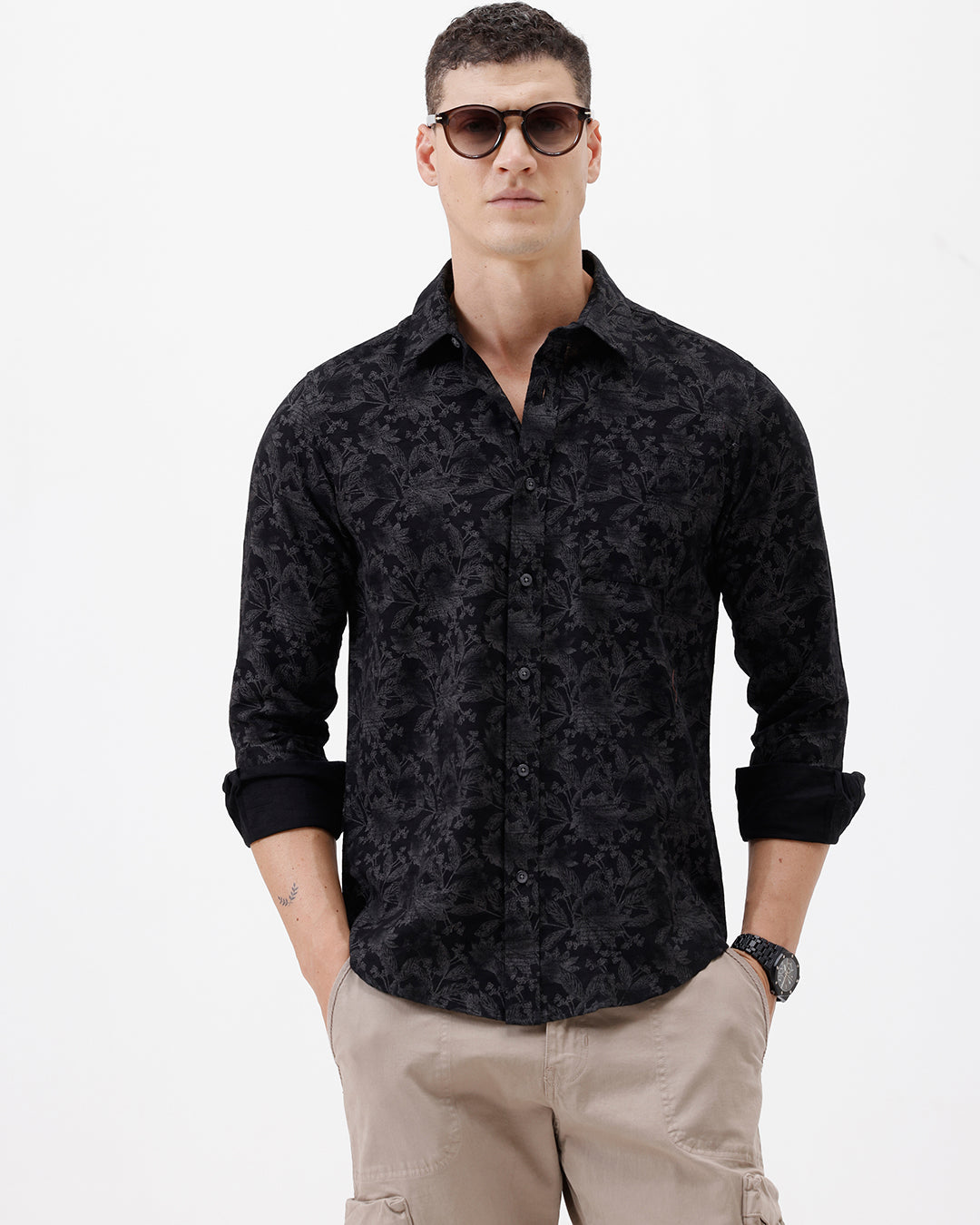 A man wearing a black long-sleeve button-up shirt with a subtle tonal floral pattern, paired with beige cargo pants and dark sunglasses, standing against a plain white background.