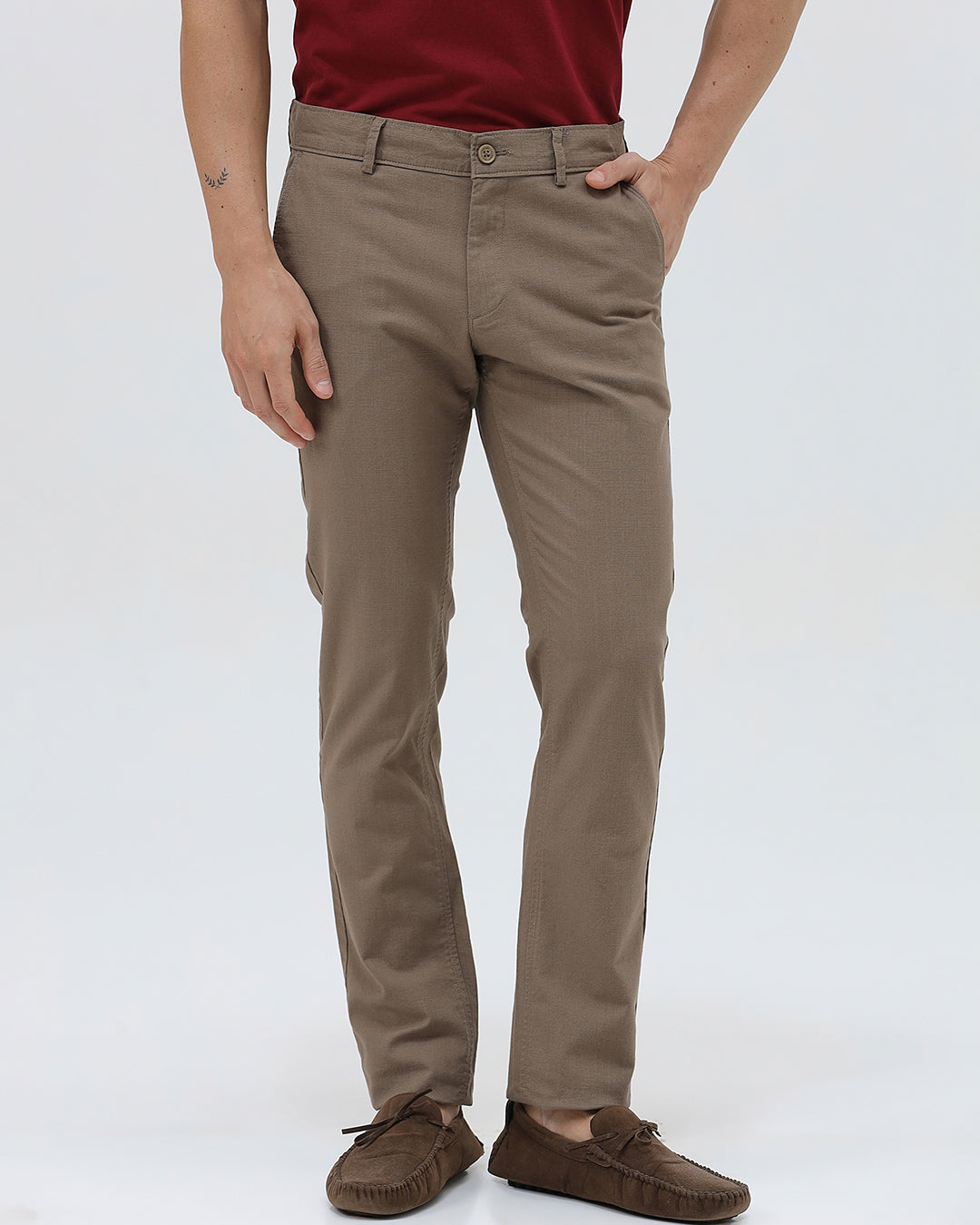 A person wearing slim-fit brown trousers, paired with a dark red shirt and brown suede moccasin shoes, shown from the waist down against a plain white studio background.