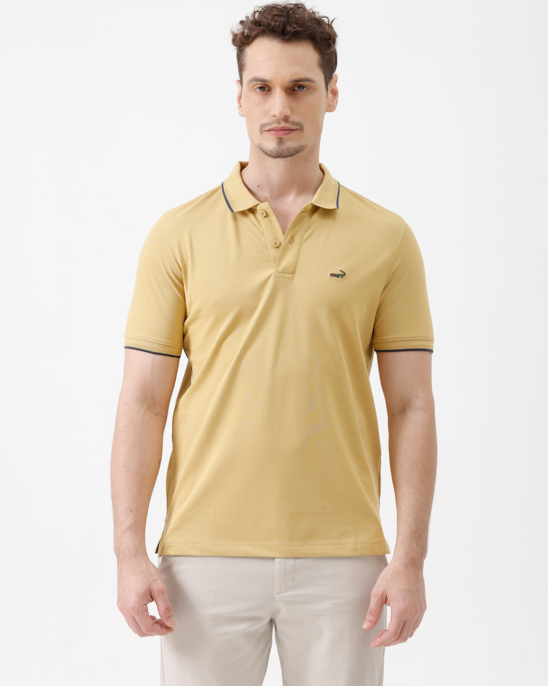 A man wearing a light yellow Lacoste polo shirt with dark piping on the collar and sleeve cuffs, paired with beige trousers, against a plain white studio background.
