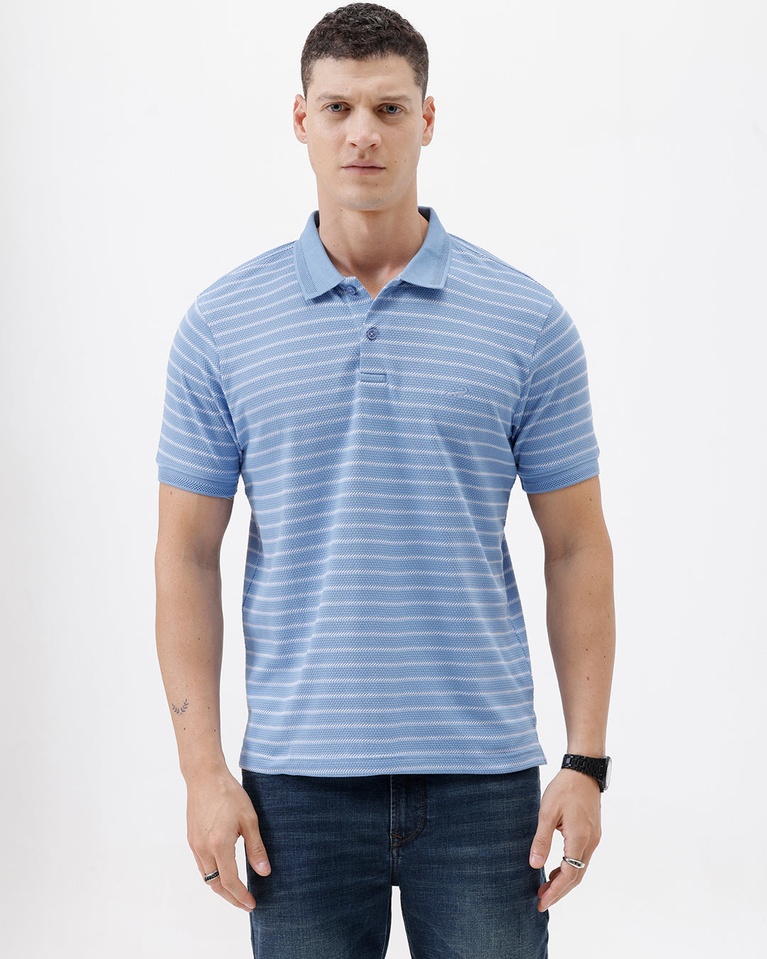 A man wearing a light blue and white striped short-sleeve polo shirt with dark blue jeans, standing against a plain light gray background.