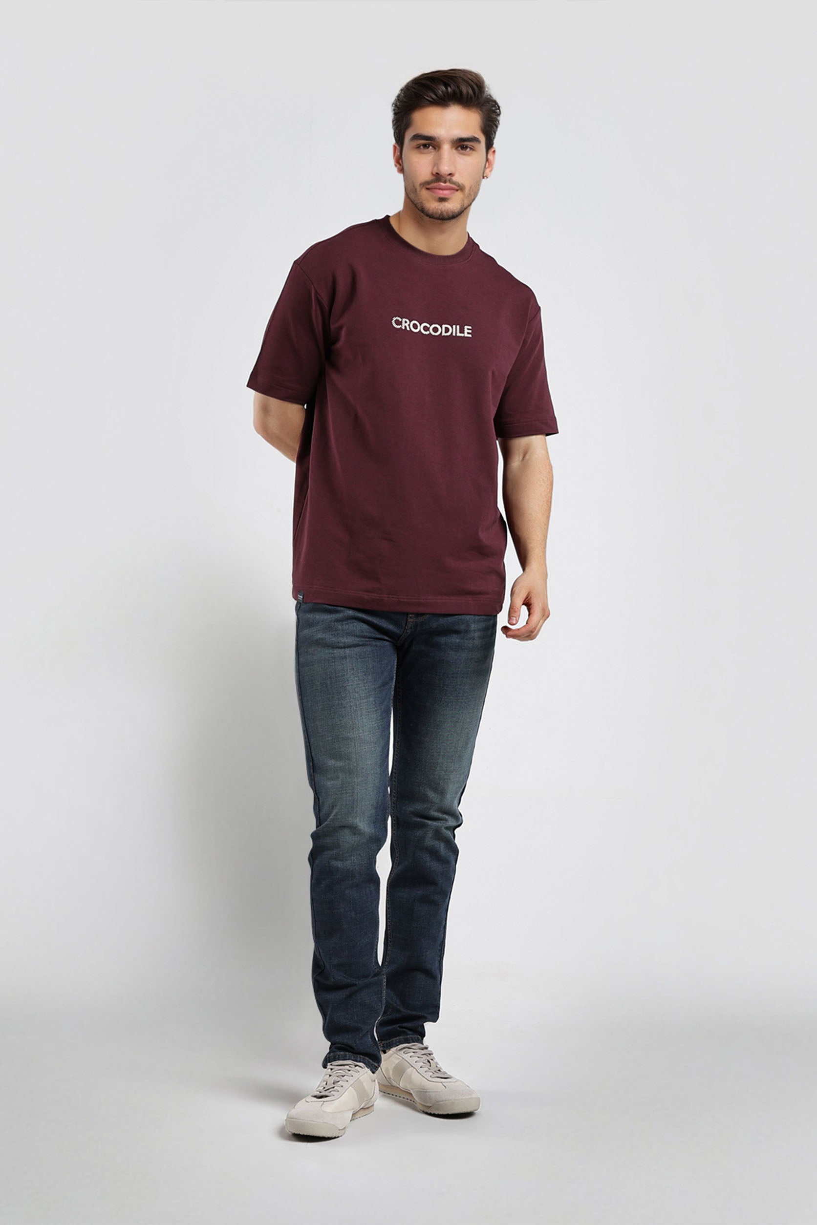 Man wearing a maroon t-shirt with a logo and dark jeans on a white background