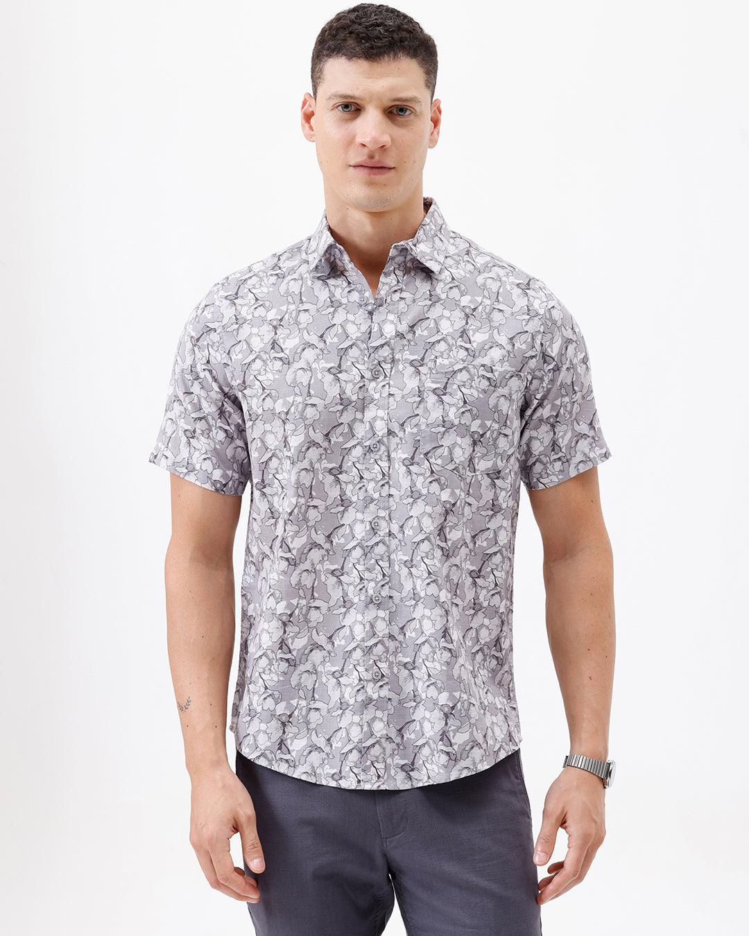 A man wearing a short-sleeve button-up shirt with a gray and white abstract pattern, paired with dark gray trousers, standing against a plain white studio background.
