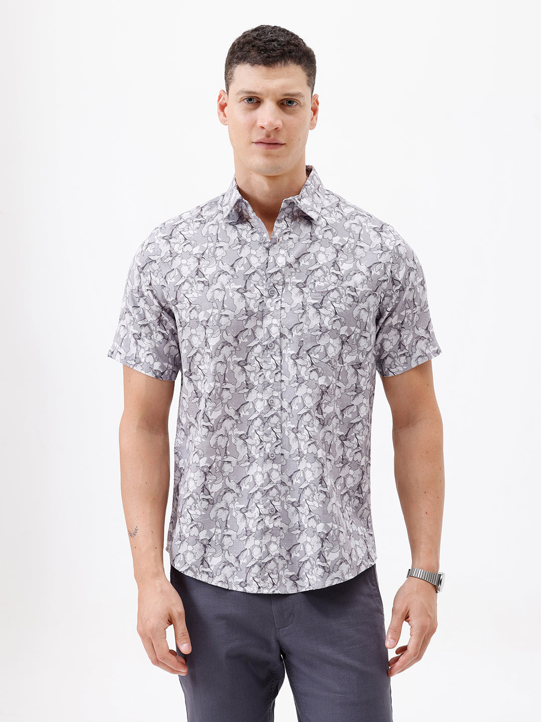 A man wearing a short-sleeve button-up shirt with a gray and white abstract pattern, paired with dark gray trousers, standing against a plain white studio background.
