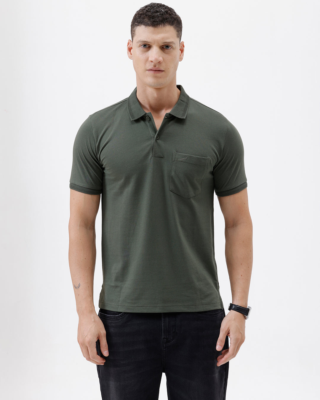 A man wearing a dark olive green short-sleeve polo shirt with a chest pocket, paired with dark jeans, standing against a plain light gray background.