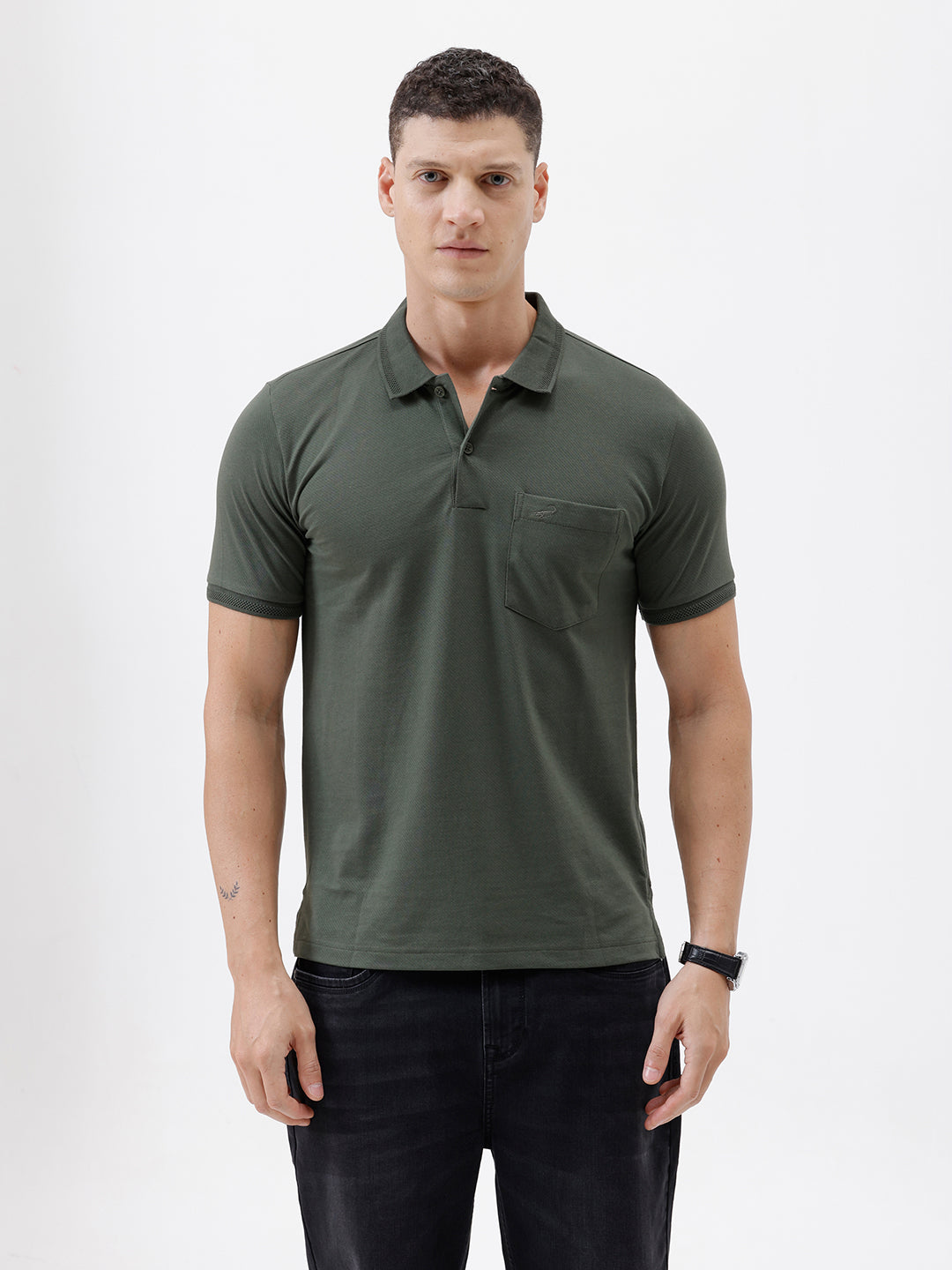 A man wearing a dark olive green short-sleeve polo shirt with a chest pocket, paired with dark jeans, standing against a plain light gray background.