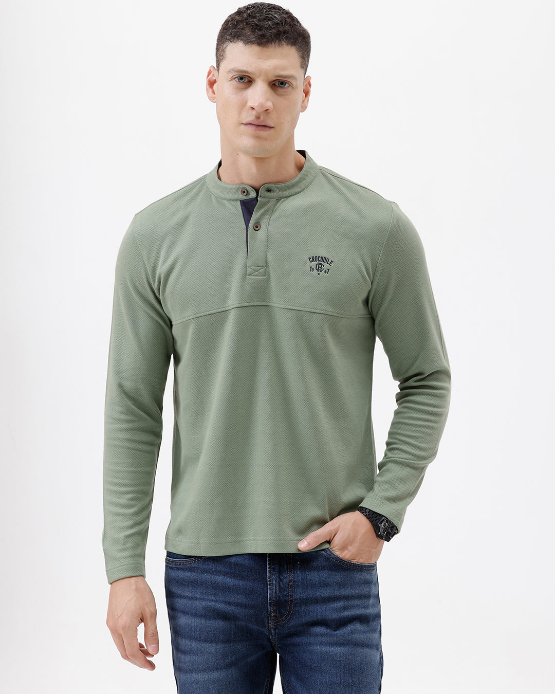 A man wearing a sage green long-sleeve henley shirt with a small embroidered logo on the left chest, paired with blue jeans, against a plain white studio background.