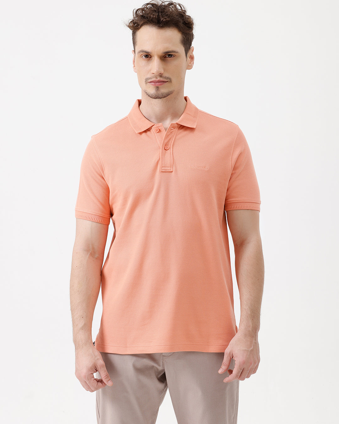 A man wearing a peach-colored short-sleeve polo shirt and beige trousers, standing against a plain white studio background.