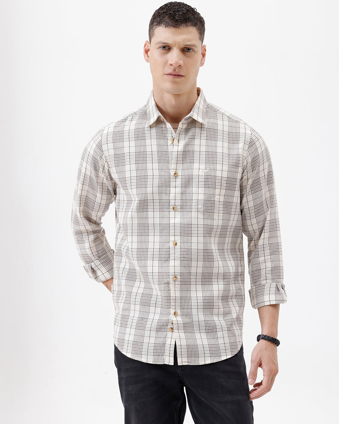 A man wearing a long-sleeve button-up plaid shirt in beige, white, and gray, paired with dark pants, standing against a plain white studio background.