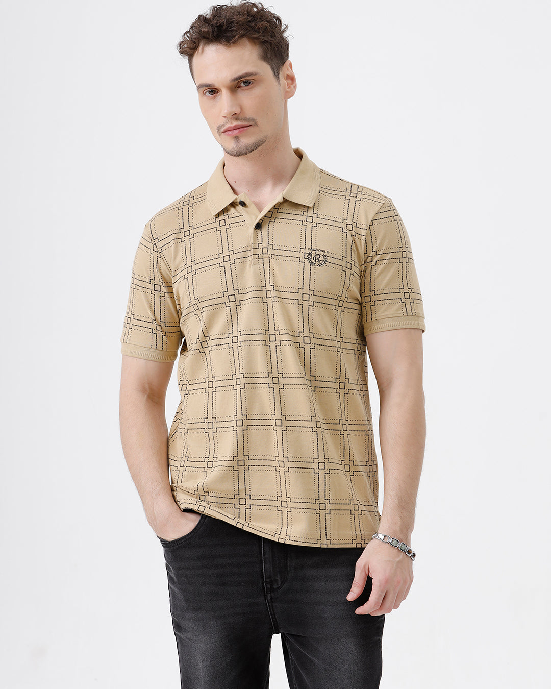 A man wearing a beige short-sleeve polo shirt with a brown grid pattern, paired with black jeans, standing against a plain white studio background.