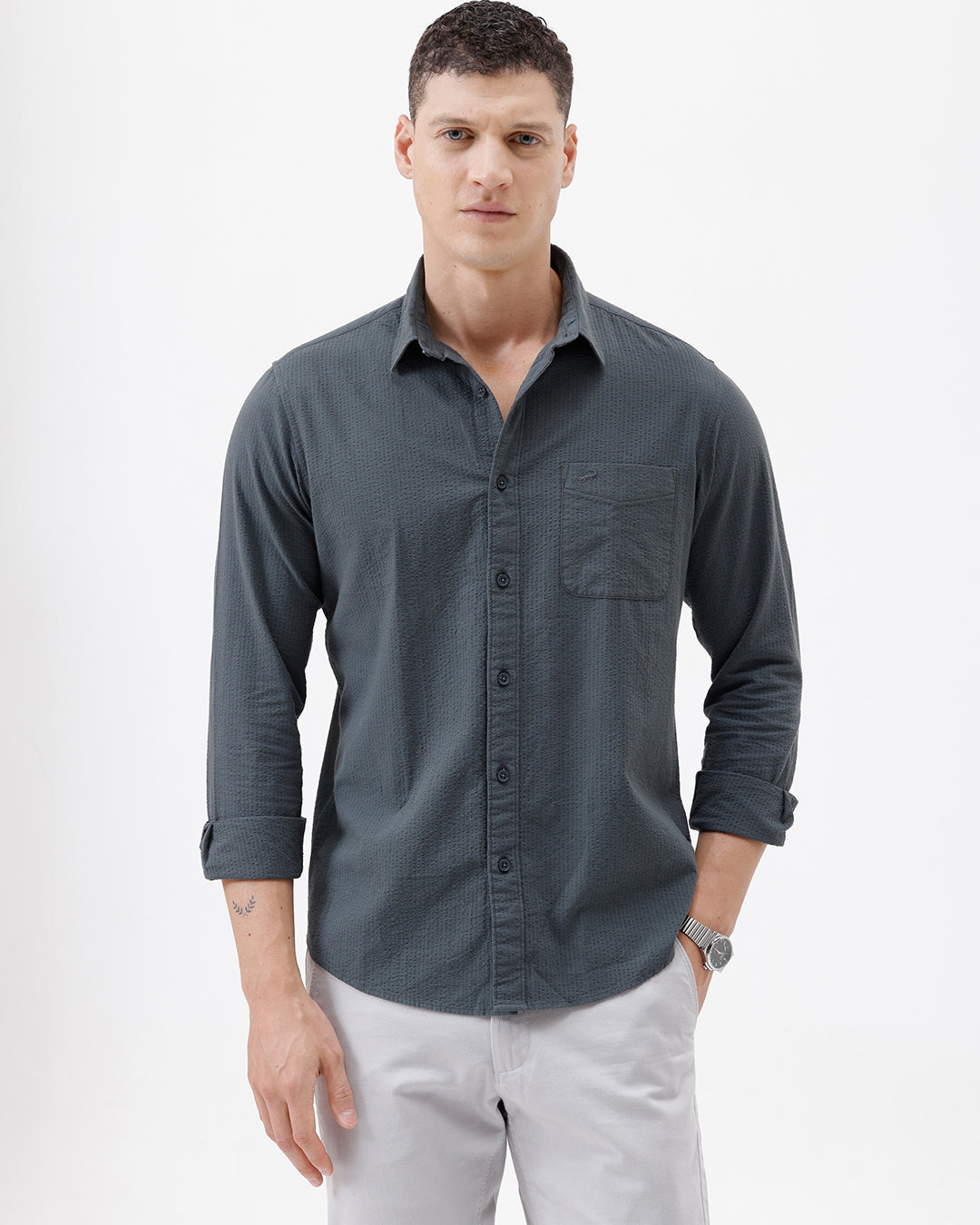 A man wearing a dark charcoal gray long-sleeve button-down shirt with a chest pocket, paired with light gray trousers, standing against a plain white studio background.