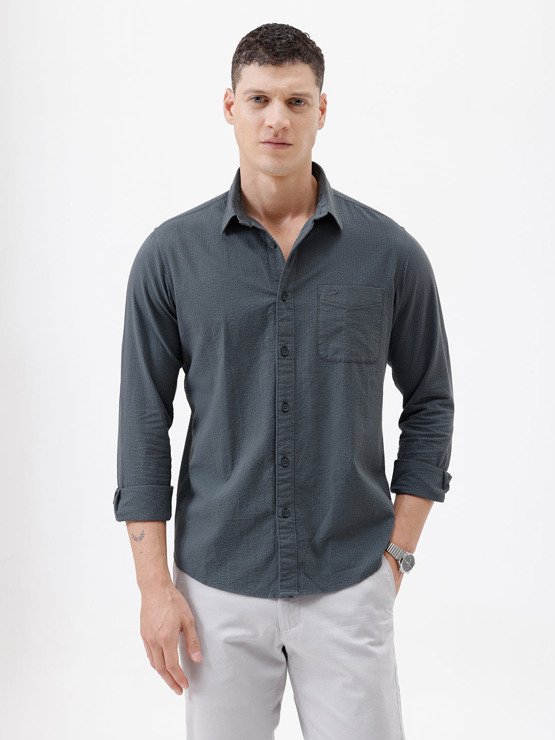 A man wearing a dark charcoal gray long-sleeve button-down shirt with a chest pocket, paired with light gray trousers, standing against a plain white studio background.