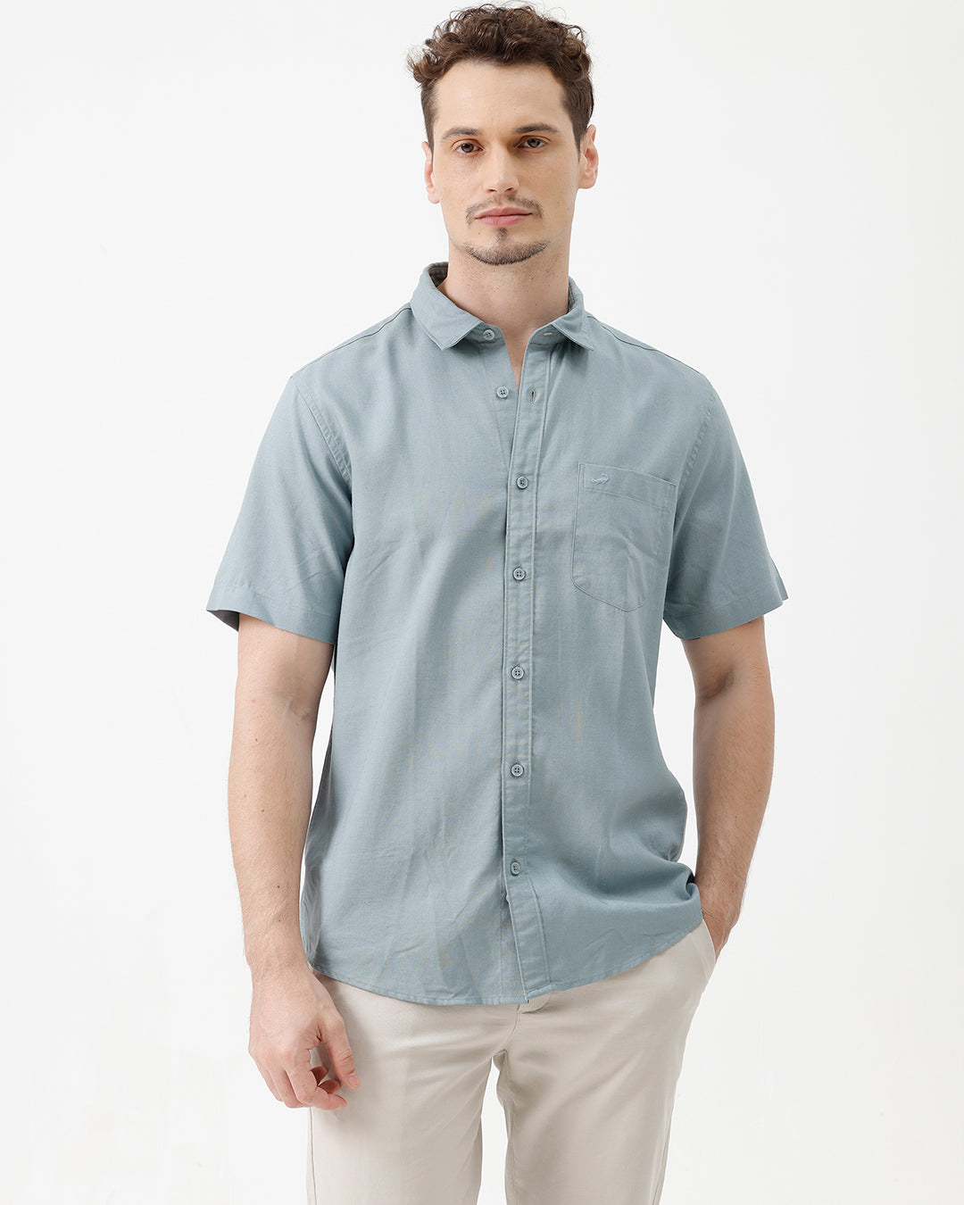 A man wearing a light blue short-sleeve button-up shirt and beige trousers, standing against a plain white studio background.