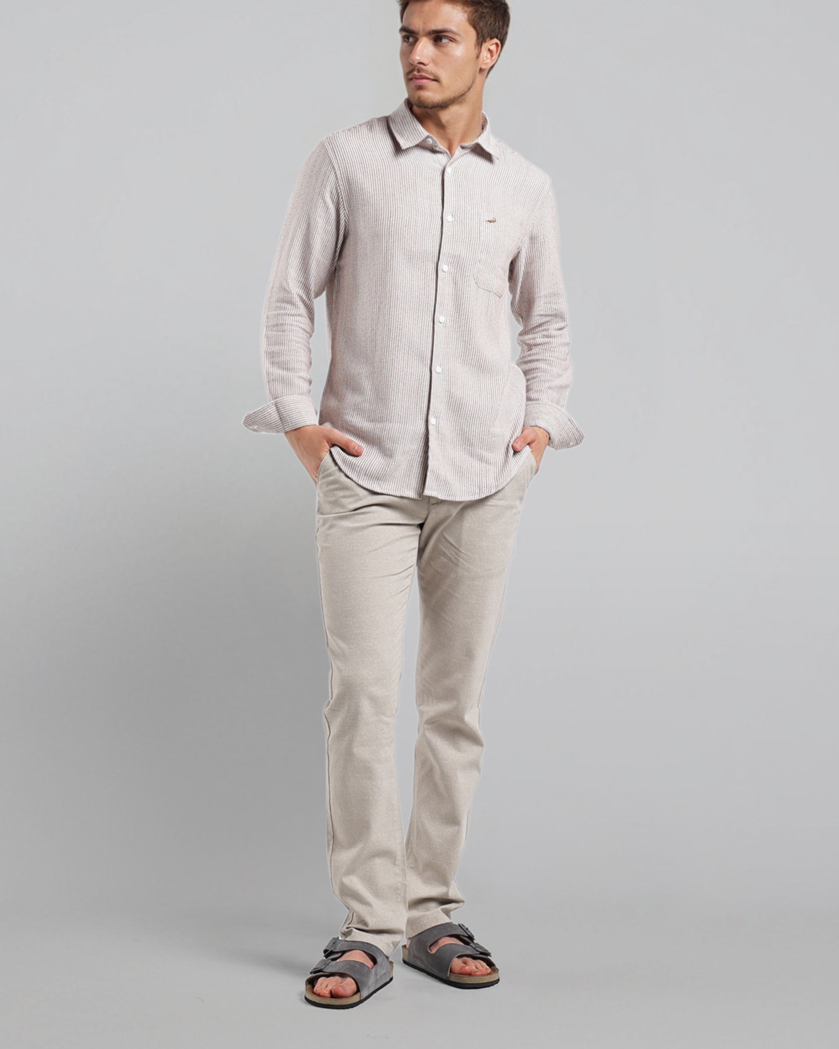 Man wearing a light gray shirt and pants with sandals on a plain background