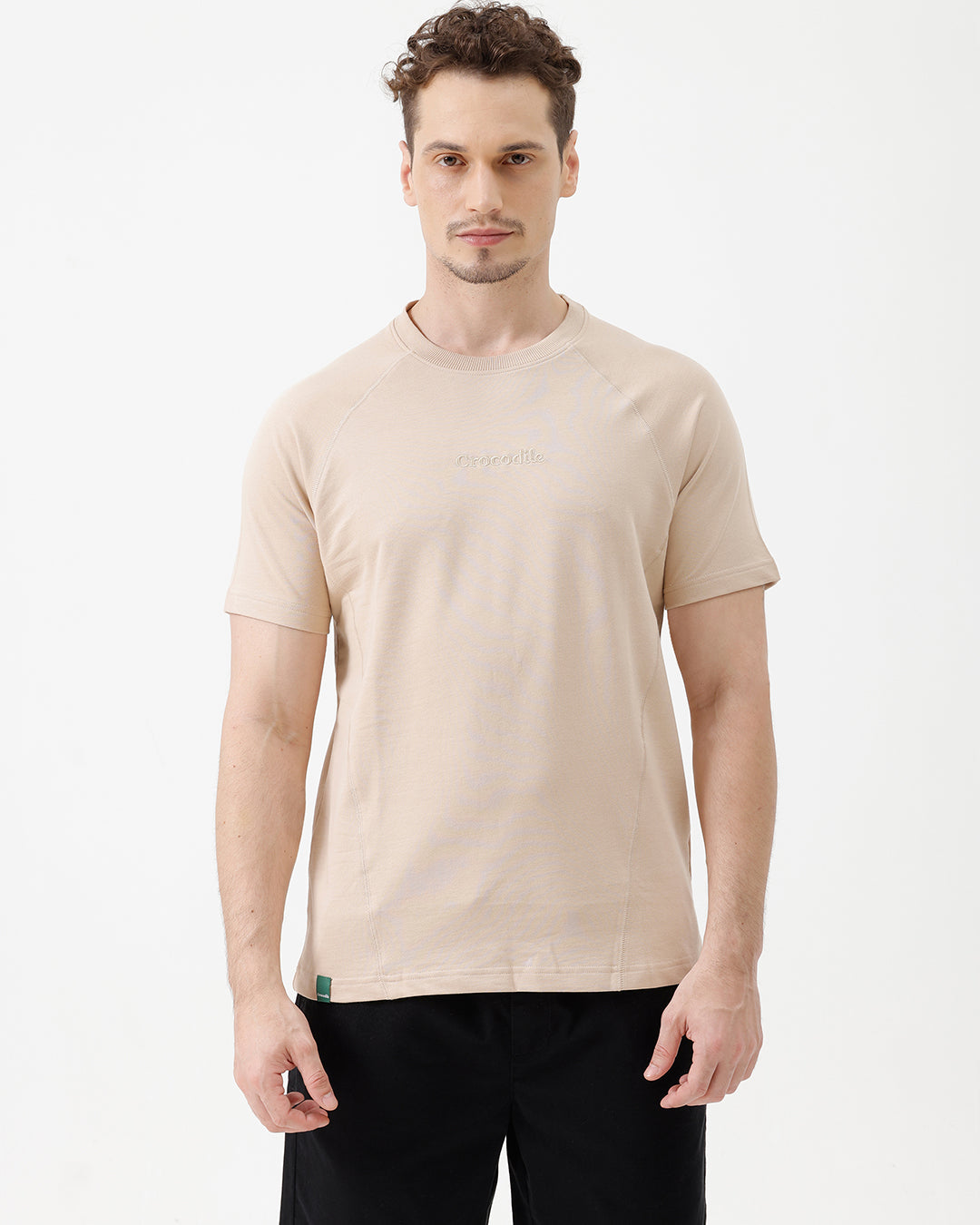 A man wearing a light beige short-sleeve t-shirt with a subtle tonal logo on the chest, paired with black pants, standing against a plain white studio background.