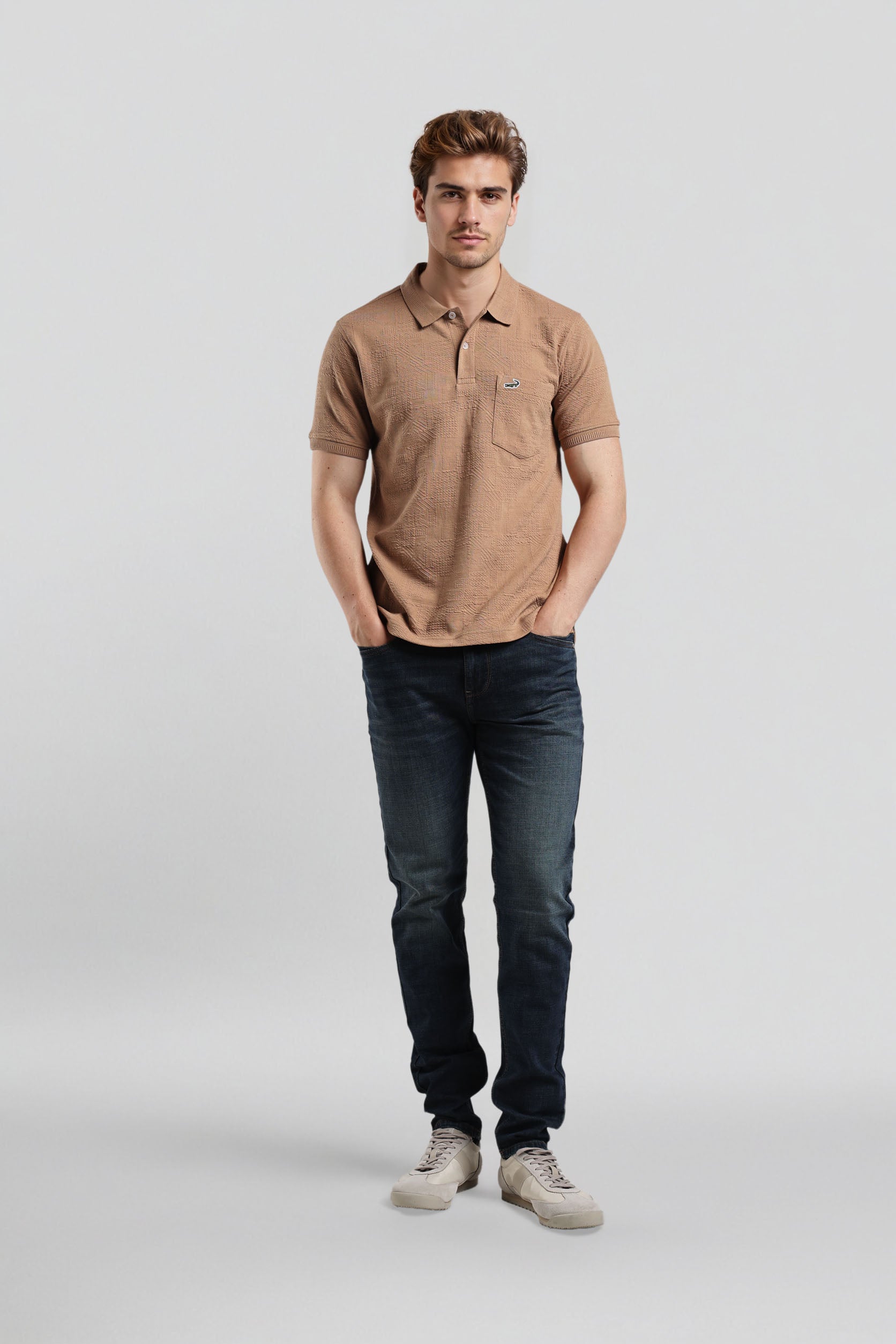 Man wearing a brown polo shirt and dark jeans on a light gray background