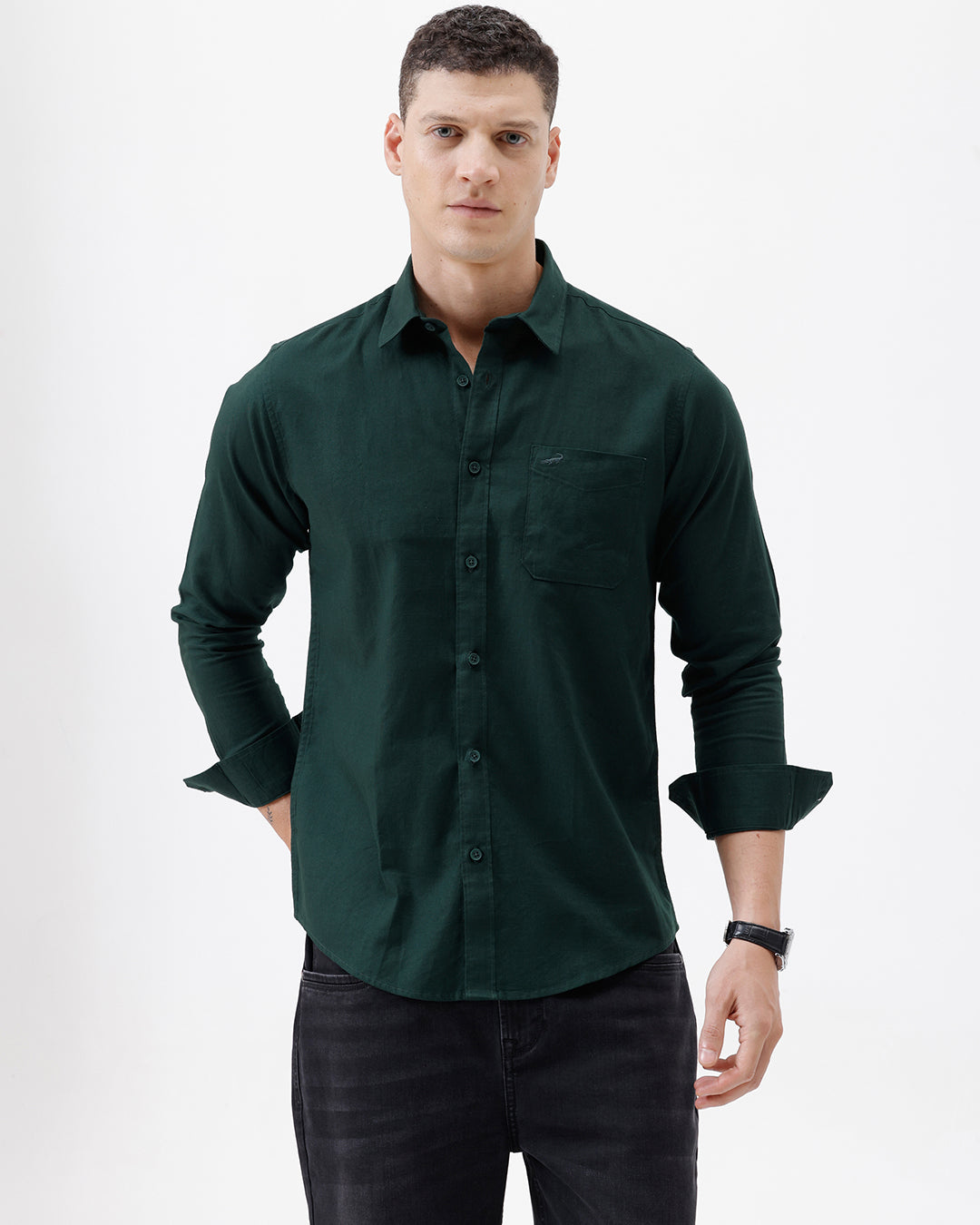 A man wearing a dark forest green long-sleeve button-down shirt with a chest pocket, paired with dark jeans, standing against a plain white studio background.