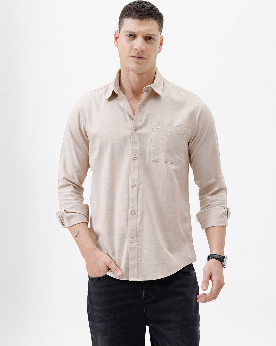 A man wearing a light beige long-sleeve button-up shirt with a chest pocket, paired with dark jeans, standing against a plain white studio background.