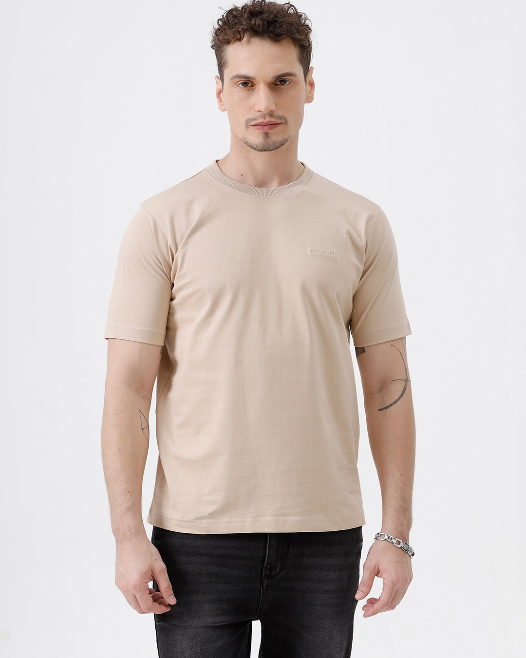 A man wearing a beige short-sleeve crewneck t-shirt with a subtle tonal logo on the left chest, paired with black jeans, standing against a light gray studio background.