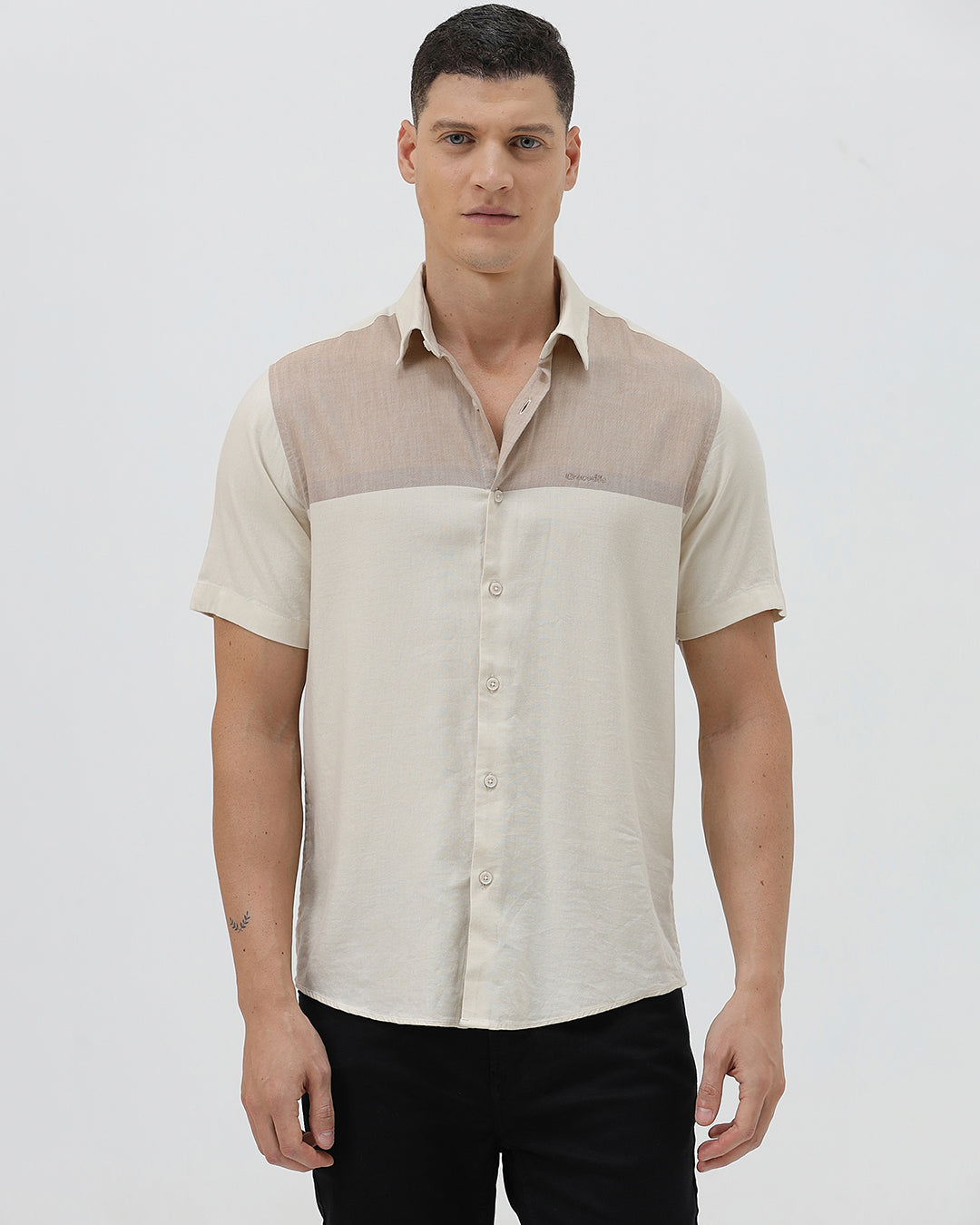 A man wearing a short-sleeve linen button-up shirt with a color-blocked design in beige and cream, paired with black pants, against a plain white studio background.