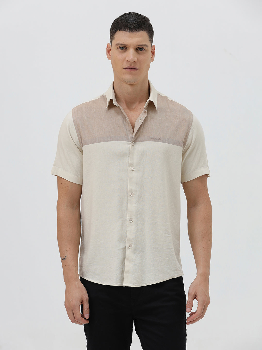 A man wearing a short-sleeve linen button-up shirt with a color-blocked design in beige and cream, paired with black pants, against a plain white studio background.