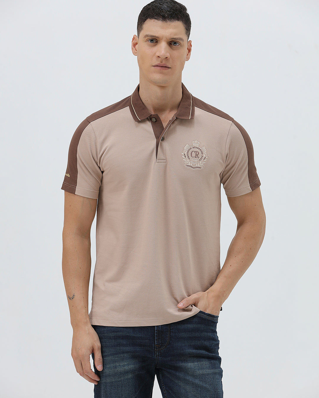 A man modeling a short-sleeve polo shirt in beige with brown shoulder panels and collar trim, paired with dark blue jeans, against a light gray studio background.