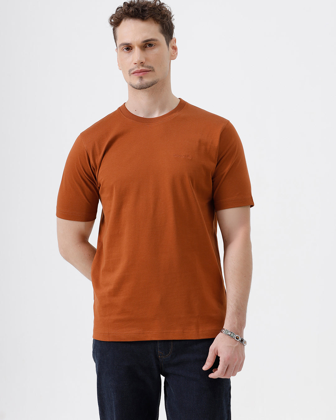 A man wearing a rust-orange crew-neck t-shirt and dark blue jeans, standing against a light gray studio background.