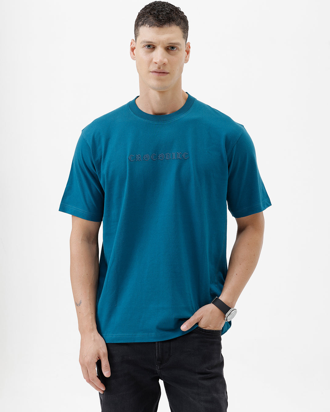 A man wearing a teal blue short-sleeve t-shirt with a subtle tonal 'GREATNESS' text graphic across the chest, paired with black pants, photographed against a clean white studio background.