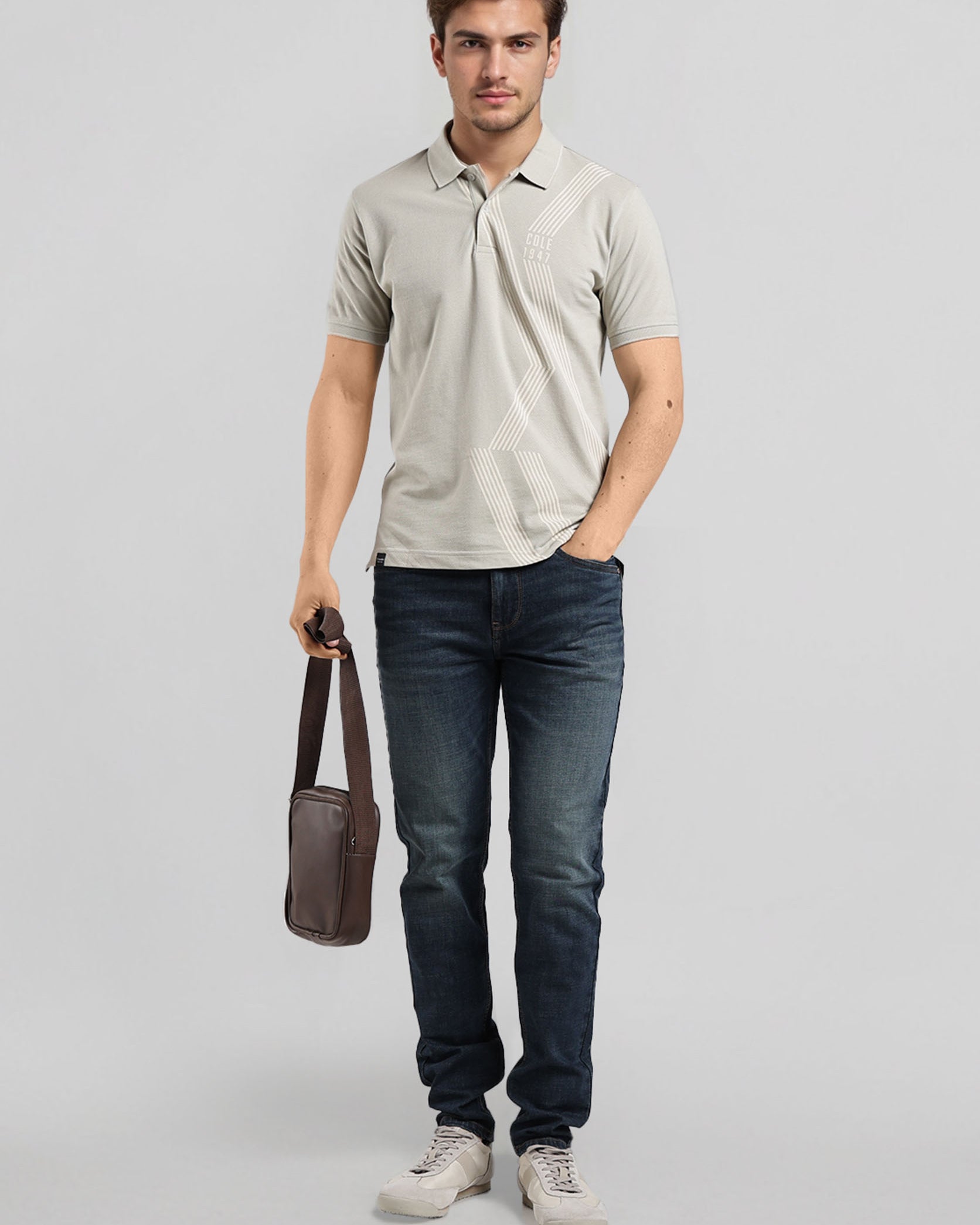 Man wearing a light gray polo shirt and dark blue jeans, holding a brown leather bag on a light gray background